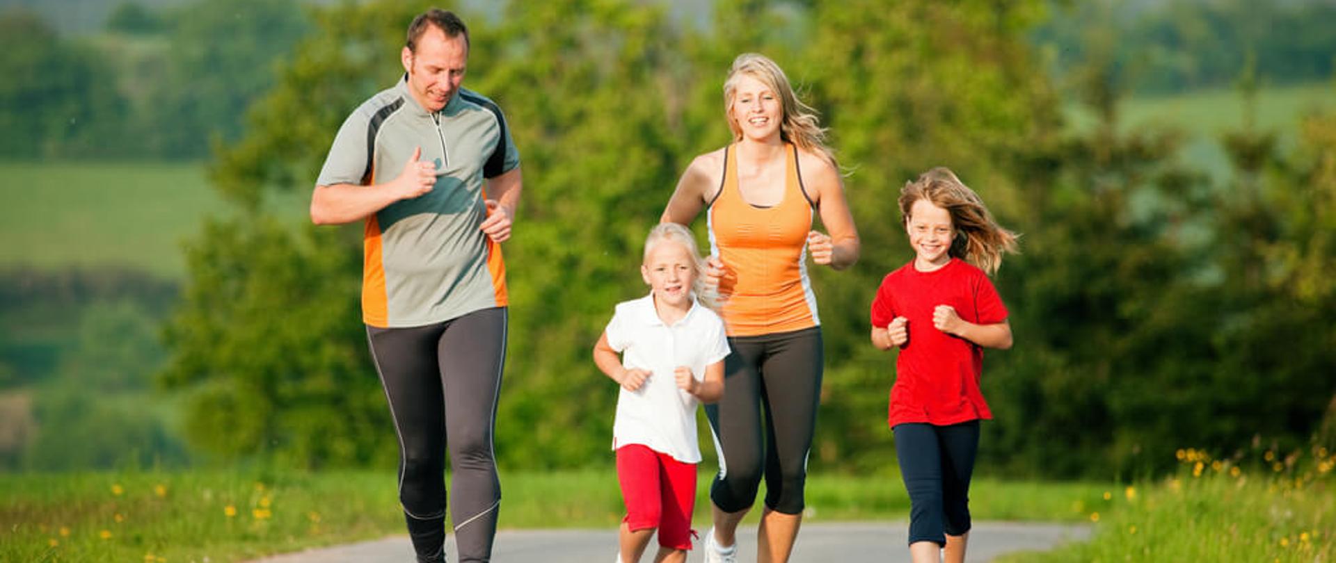 na pierwszym planie dwie osoby dorosłe oraz dwoje dzieci. Osoby dorosłe w wieku około 35 lat, dzieci w wieku około 5 lat. wszyscy biegną po ścieżce asfaltowej biegnącej wśród łąk. wszyscy ubrani w stroje sportowe 