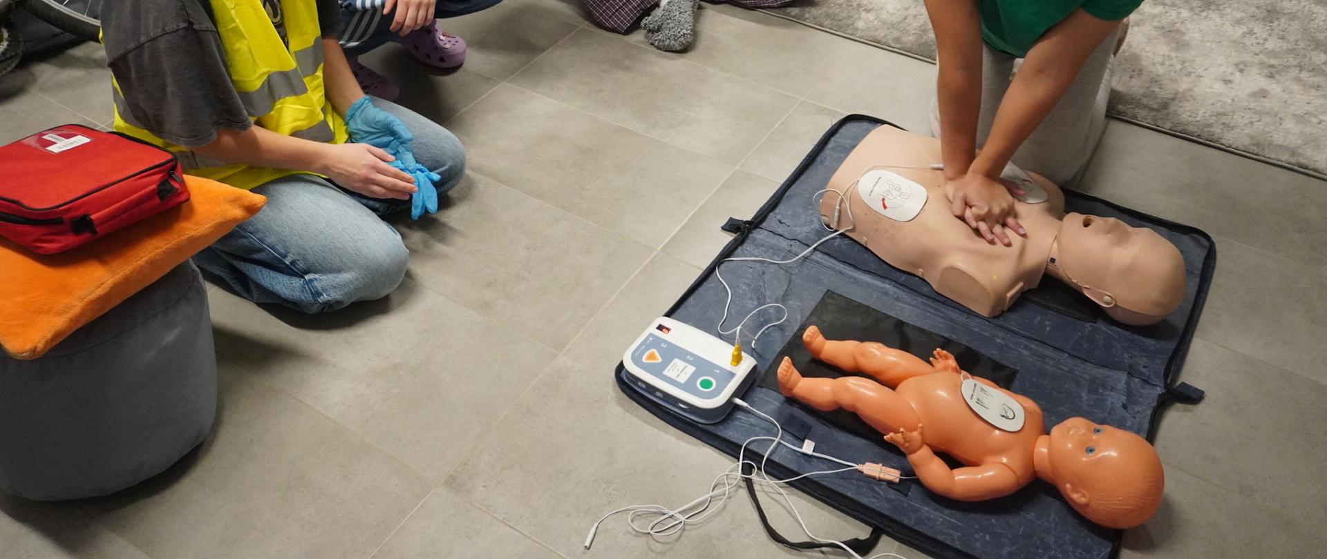 Wychowanka wykonuje uciśnięcia klatki piersiowej. Obok leży defibrylator.