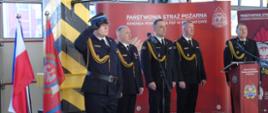 Na zdjęciu w mundurach wyjściowych komendanci oraz prowadzący uroczystość w tle baner straży oraz flagi