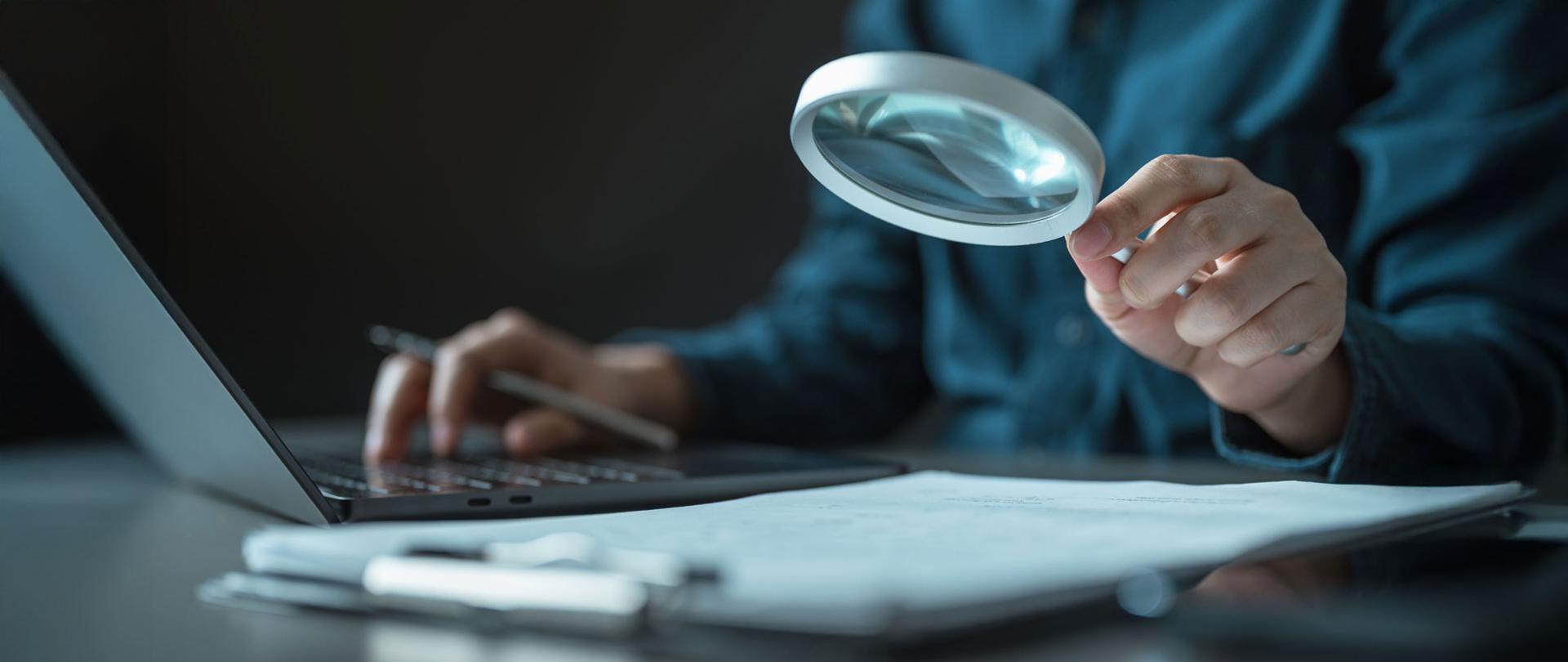 Osoba siedząca przy biurku trzyma lupę nad dokumentami leżącymi obok otwartego laptopa.