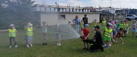 Zdjęcie przedstawia strażaka oraz dzieci ubrane w kamizelki odblaskowe. Oboje leja wodę z prądownicy wodnej.