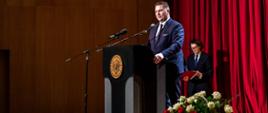 Jubileusz-20-lecia Uniwersytetu Rzeszowskiego z udziałem Ministra Edukacji i Nauki Przemysława Czarnka minister przemawia do mikrofonu. w tle czerwona załona, z prawej stronu ministra kosz z biało czerwonymi różami