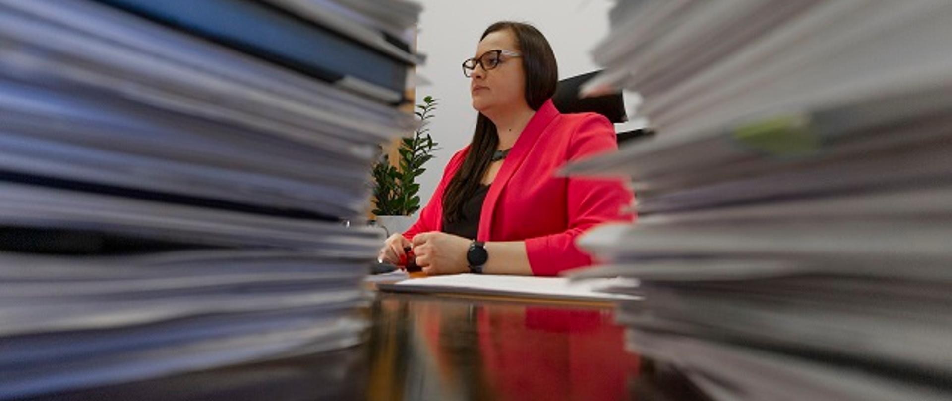 Zdjęcie minister Małgorzaty Jarosińskiej-Jedynek za biurkiem. Kadr pokazuje Minister między dwoma stosami dokumentów.