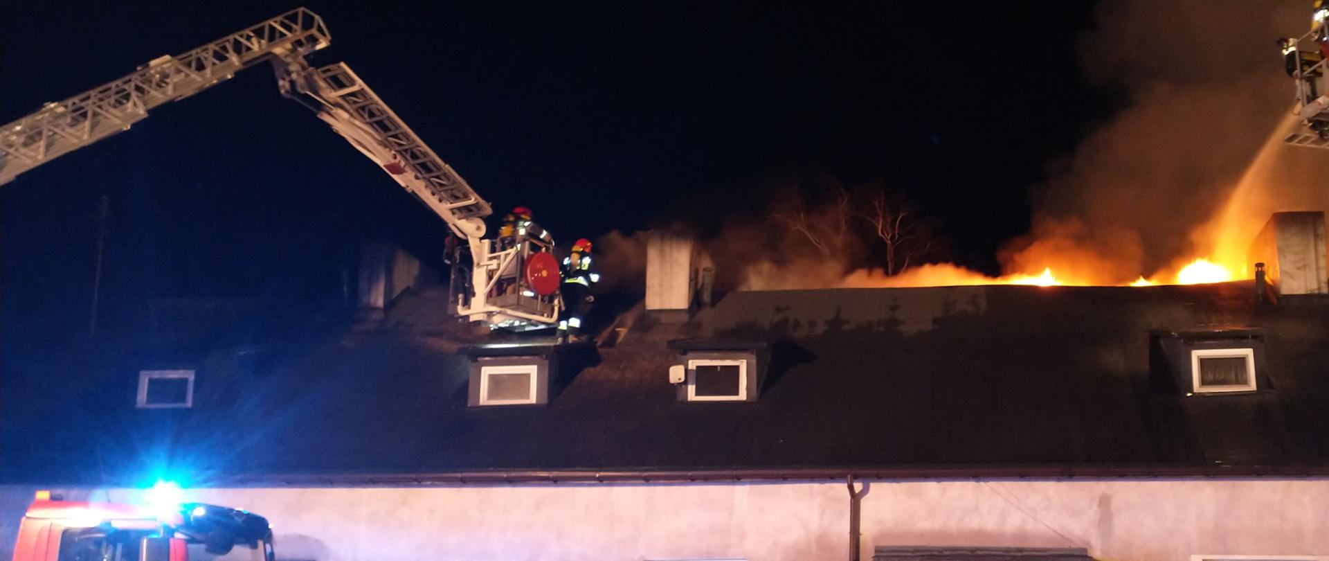Na zdjęciu widać palący się parterowy z poddaszem użytkowym budynek mieszkalny wielorodzinny. Z podnośników strażacy leją wodę na dach, na którym widać płomienie ognia. Przed budynkiem stoi strażacki podnośnik oraz ciemny samochód osobowy. Jest ciemno.