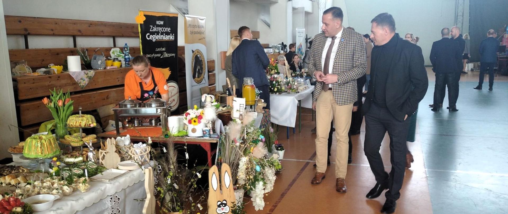 dwóch mężczyzn ogląda stoisko udekorowane palmami wielkanocnymi, drewnianymi zającami wielkanocnymi i ciastami, babkami wielkanocnymi i jajkami faszerowanymi