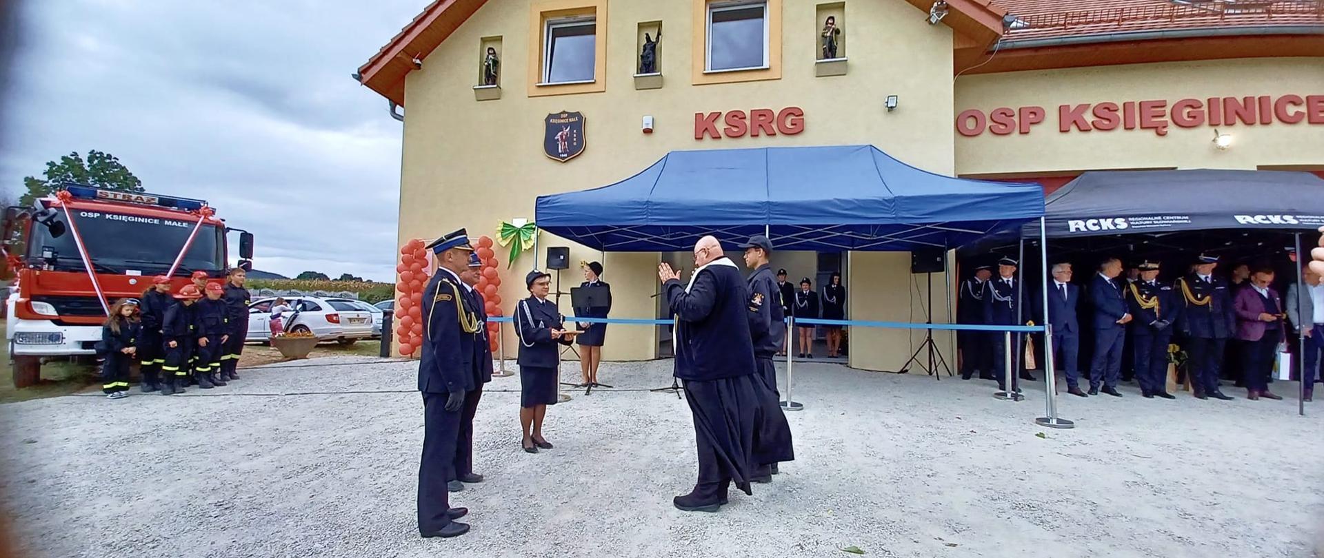 Kilkanaście osób na uroczystości otwarcia nowej remizy. W tle widoczna elewacja budynku i samochód ratowniczo - gaśniczy