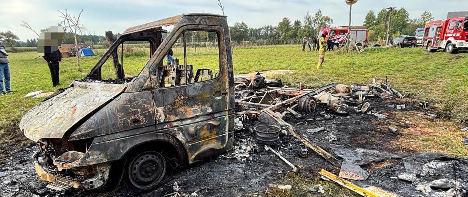 Zwęglony pojazd dostawczy po pożarze, znajdujący się na polu. W tle widoczne są zastępy straży pożarnej oraz strażacy prowadzący działania.