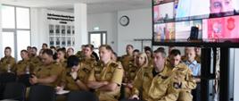 Funkcjonariusze Państwowej Straży Pożarnej siedzą obok siebie na krzesłach podczas szkolenia na ekranie telewizora wyświetlane są połączenia z poszczególnymi komendami Państwowej Straży Pożarnej.