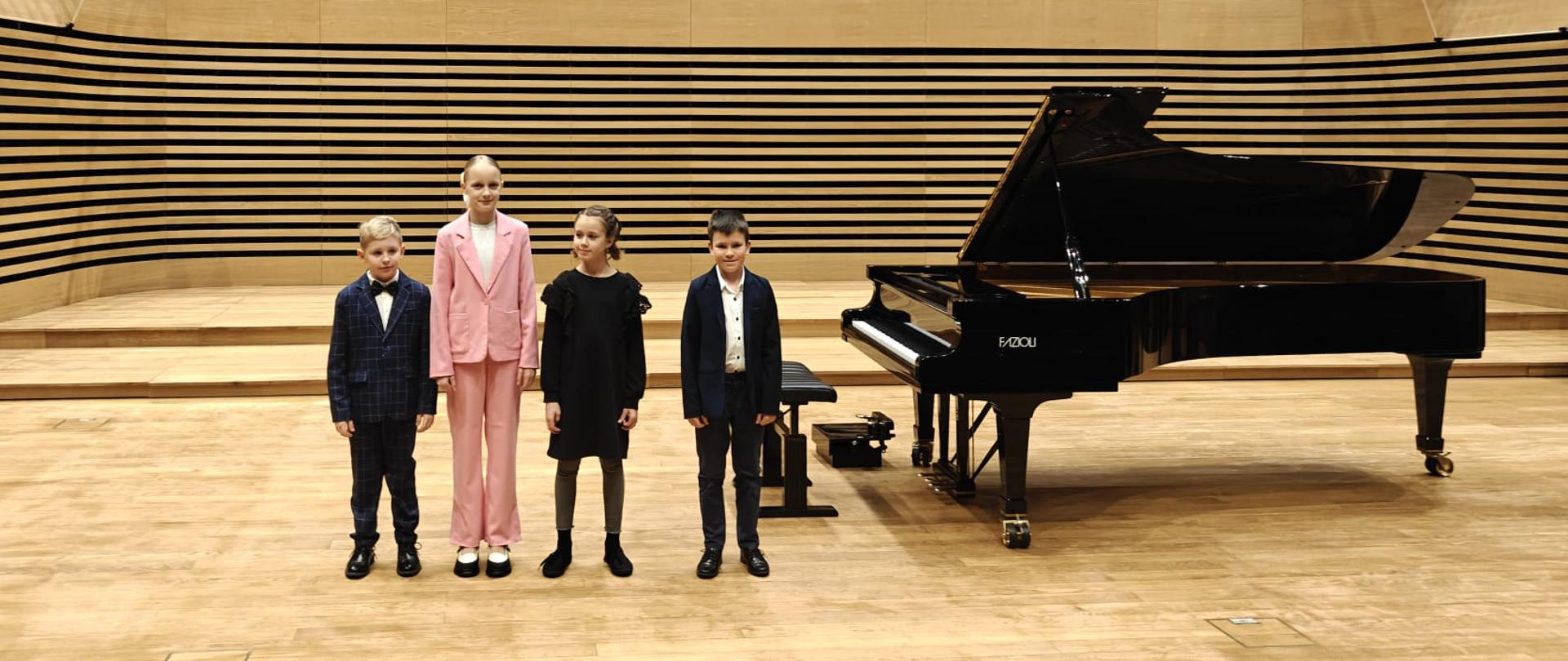 Dwie dziewczynki i dwóch chłopców stoi na estradzie przy fortepianie.