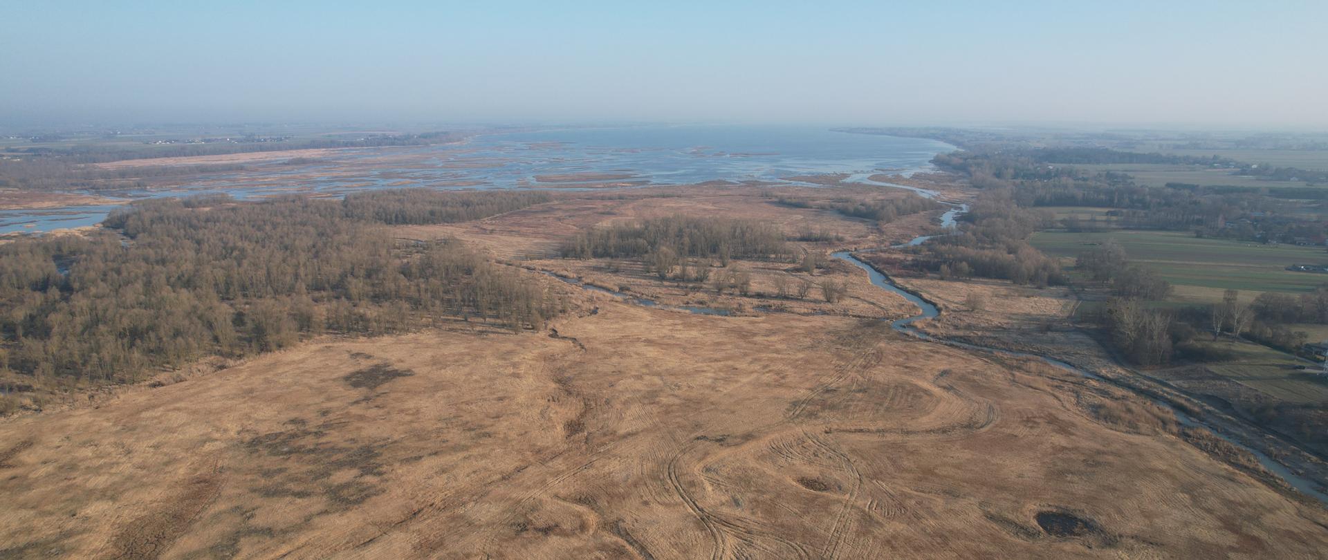 Widok z lotu ptaka na rozległy krajobraz doliny rzecznej. Przez środek kadru meandruje rzeka, tworząc liczne zakola i rozlewiska. Wokół widoczne są łąki i tereny podmokłe w odcieniach brązu, z kępami drzew i zarośli.