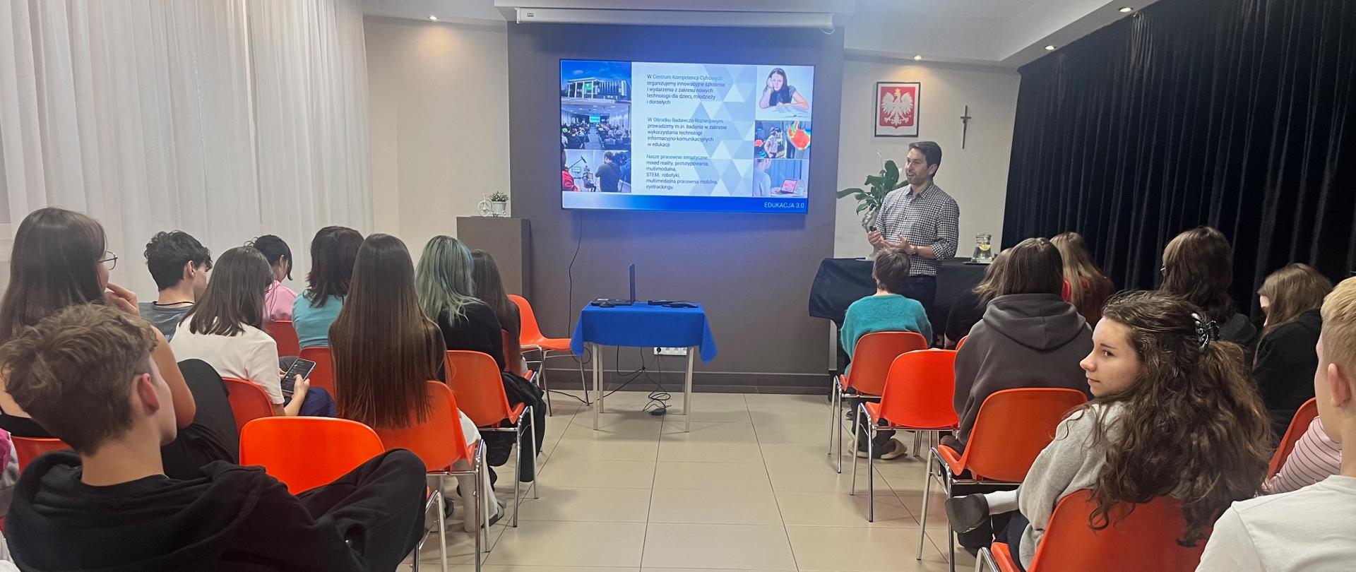 W pierwszym planie widać od tyłu kilkunastu młodych ludzi siedzących na pomarańczowych krzesłach ustawionych w dwóch kolumnach. Na ścianie na wprost wisi duży ekran telewizyjny, a na nim wyświetla się kilka zdjęć i tekst. Przed ekranem stoi stolik z niebieskim obrusem i laptopem. Po prawej stronie stoi mężczyzna zwrócony w stronę osób siedzących przed nim. Na ścianie za nim wisi krzyż i godło Polski. Na ścianie po prawej są czarne zasłony, a po lewej białe.