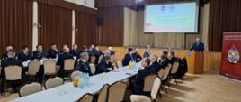 Strażacy ubrani w granatowe mundury galowe, oraz pisakowe mundury siedzą przy stolikach, w tyle rolap z logiem Państwowej Straży Pożarnej na tylnej ścianie kotary oraz ekran z wyświetloną prezentacją. Przy mównicy stoi starosta ubrany w garnitur, przed sobą ma mikrofon