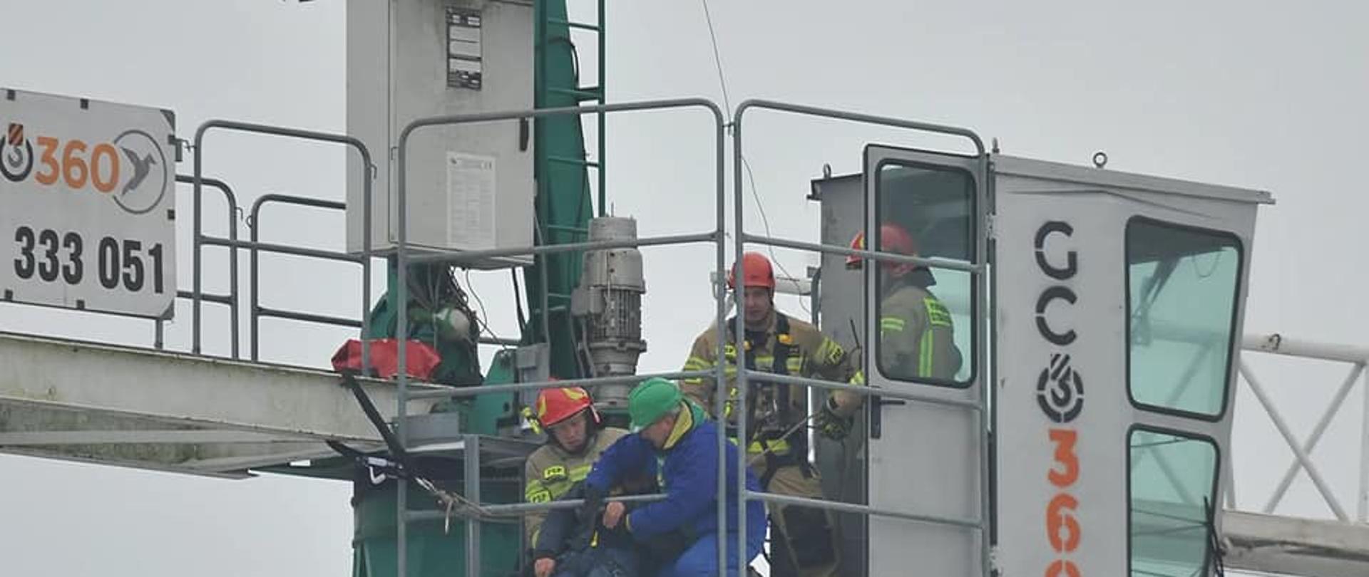 Na zdjęciu widać podest oraz kabinę żurawia wieżowego. W kabinie oraz na podeście znajdują się strażacy w ubraniach specjalnych oraz pracownik firmy budowlanej, którzy przy użyciu technik alpinistycznych przygotowują się do spuszczenia poszkodowanego z podestu.