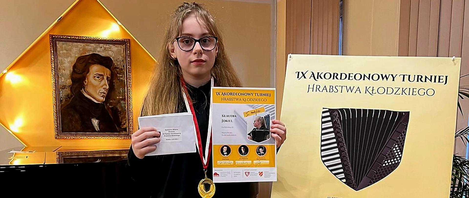 Na zdjęciu uczennica trzymająca w jednej ręce dyplom, a w drugiej kopertę, stojąca obok sztalugii z plakatem konkursu. W tle widoczny fortepian i w ścianie - we wnęce w kształcenia rombu - na złotym tle podświetlony obraz Fryderyka Chopina. 