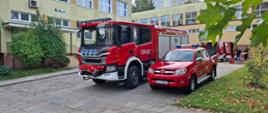 Dwa samochody strażackie zaparkowane pod szkołą. W tle widać busa strażackiego.