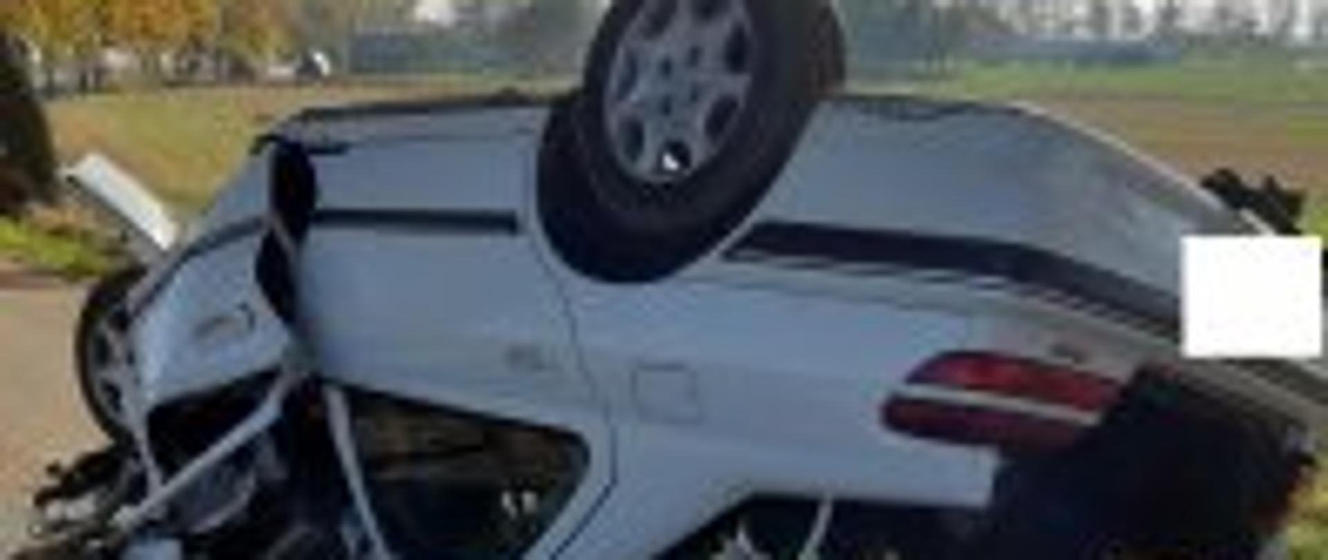 Biały samochód osobowy znajdujący się na dachu na drodze asfaltowej. Pojazd ma rozbity przód oraz otwartą tylna klapę bagażnika.