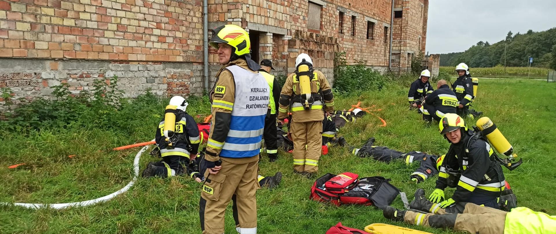 Zdjęcie przedstawia ćwiczących strażaków przed blokiem z czerwonej cegły. W centrum zdjęcia znajduje się Kierujący Działaniem Ratowniczym.