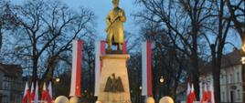 Narodowe Święto Niepodległości. Zdjęcie przedstawia pomnik Żołnierza Polskiego. Żołnierz w płaszczu i czapce rogatywce, trzymający przed sobą karabin, stoi na cokole, na którym umieszczony jest orzeł. Za pomnikiem, na pionowych masztach zawieszone są 4 flagi biało-czerwone. Z kolei z prawej i lewej strony pomnika ustawione są dwa stojaki z flagami biało-czerwonymi. W tle budynki i drzewa.