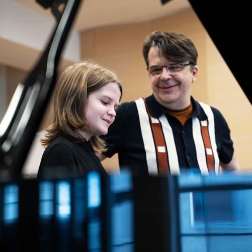 zdjęcie przedstawia młodszego uczestnika warsztatów we wspólnym zdjęciu z wykładowcą przy fortepianie na scenie sali koncertowej, ujęcie z bliska