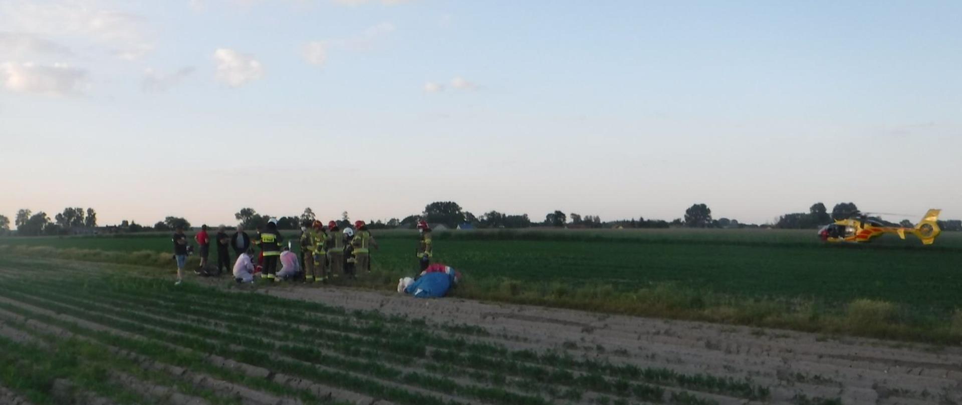 Na zdjęciu widać strażaków oraz załogę karetki pogotowia, którzy udzielają, na polu, pomocy paralotniarzowi. Obok stoją osoby postronne. W tle widać żółto-czerwony śmigłowiec Lotniczego Pogotowia Ratunkowego oraz zabudowania. Jest widno.