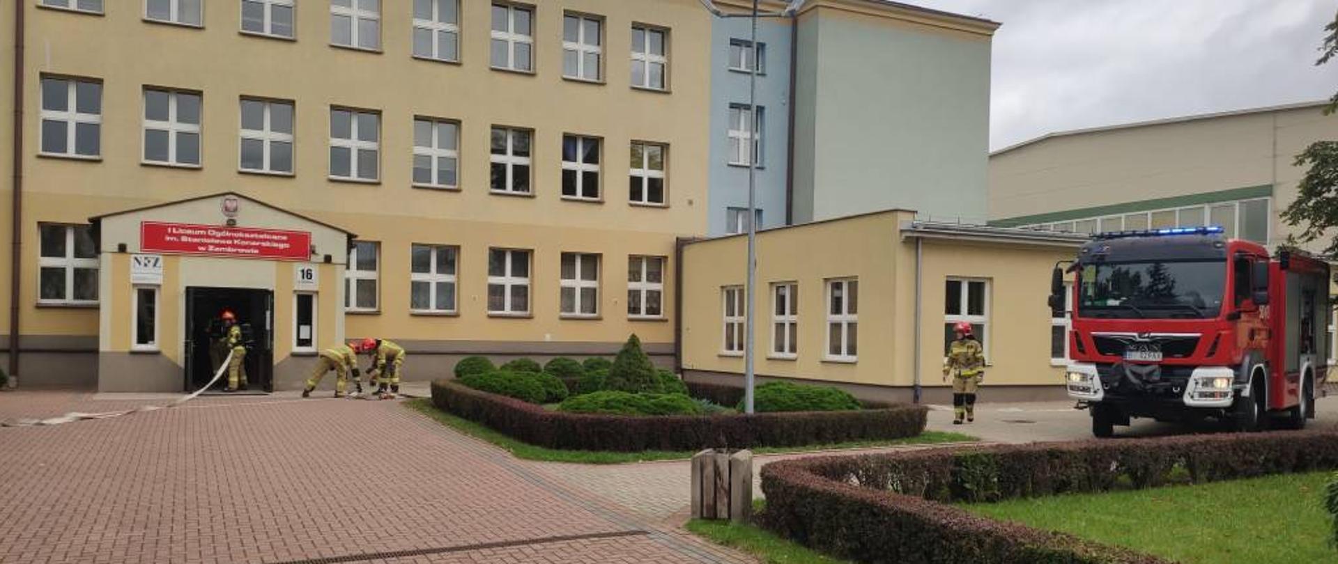 Zdjęcie przedstawia strażaków w ubraniach specjalnych podczas wchodzenia do budynku szkoły z linią gaśniczą w celu ugaszenia pozorowanego w ramach próbnej ewakuacji pożaru w jednym z pomieszczeń szkoły. Na zdjęciu widać część budynku szkoły oraz średni samochód ratowniczo-gaśniczy stojący na placu przed szkołą z włączonymi światłami mijania i niebieskimi światłami błyskowymi. Zdjęcie w pochmurny dzień wykonano przed budynkiem I Liceum Ogólnokształcącego w Zambrowie. 