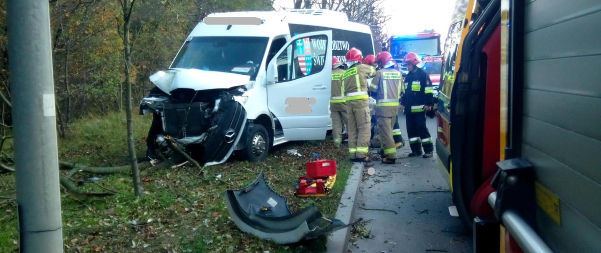 Zdjęcie przedstawia ulicę na której doszło do wypadku komunikacyjnego. Na poboczu stoi uszkodzony samochód, przed nim leżą oderwane fragmenty auta, a przy pojeździe znajduje się grupa strażaków w czasie działań. Po prawej stronie, na jezdni stoją samochody pożarnicze.