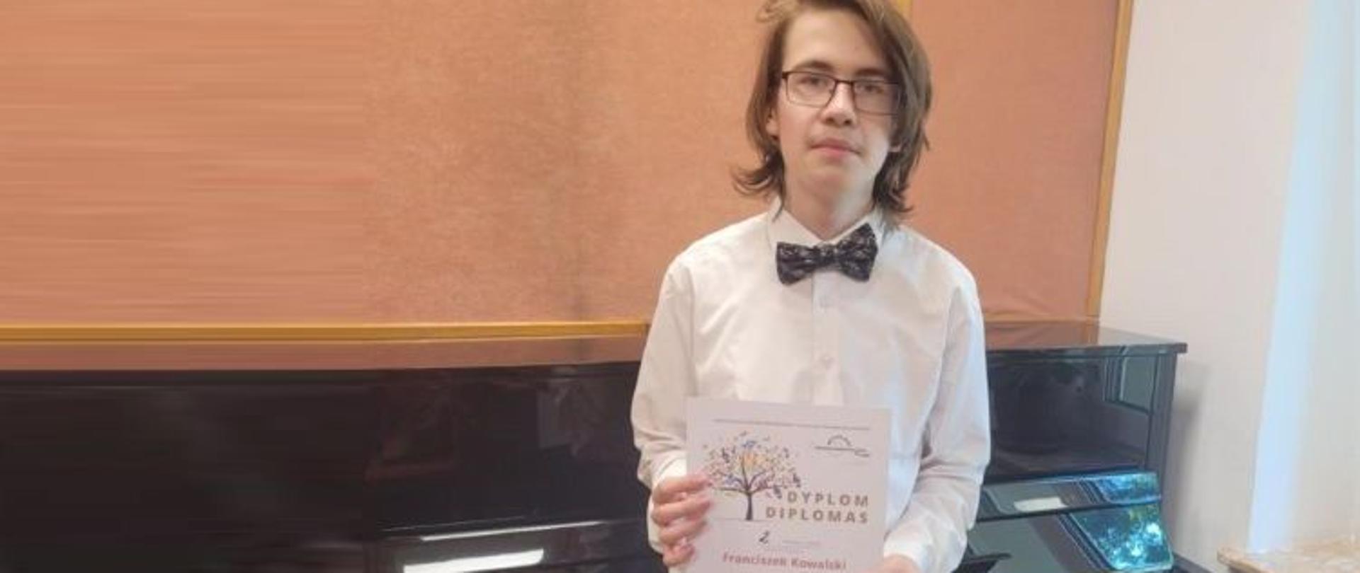 Zdjęcie kolorowe. Młody mężczyzna stojący obok fortepianu trzyma dyplom. Mężczyzna jest w białej koszuli i muszce. Fortepian jest czarny, a na nim nuty. Z prawej strony widać kawałek okna i kaloryfer.