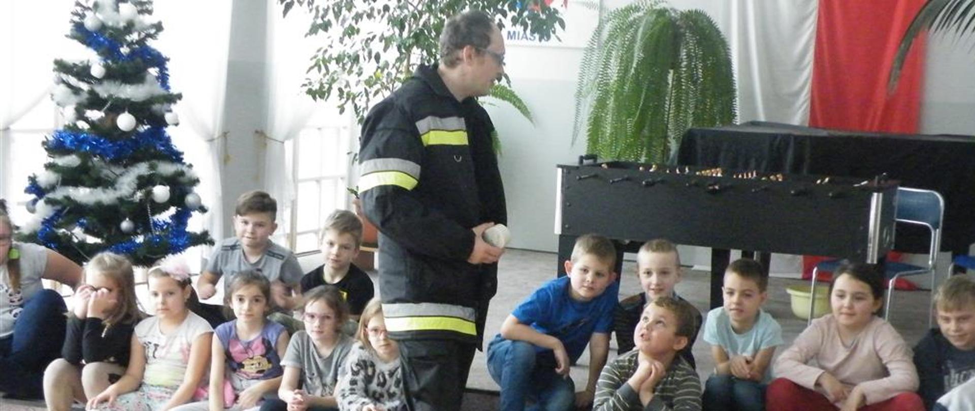 Grupa dzieci siedzi na schodkach w świetlicy szkolnej, przed nimi stoi strażak trzymający w ręku czujkę dymu, o której opowiada.