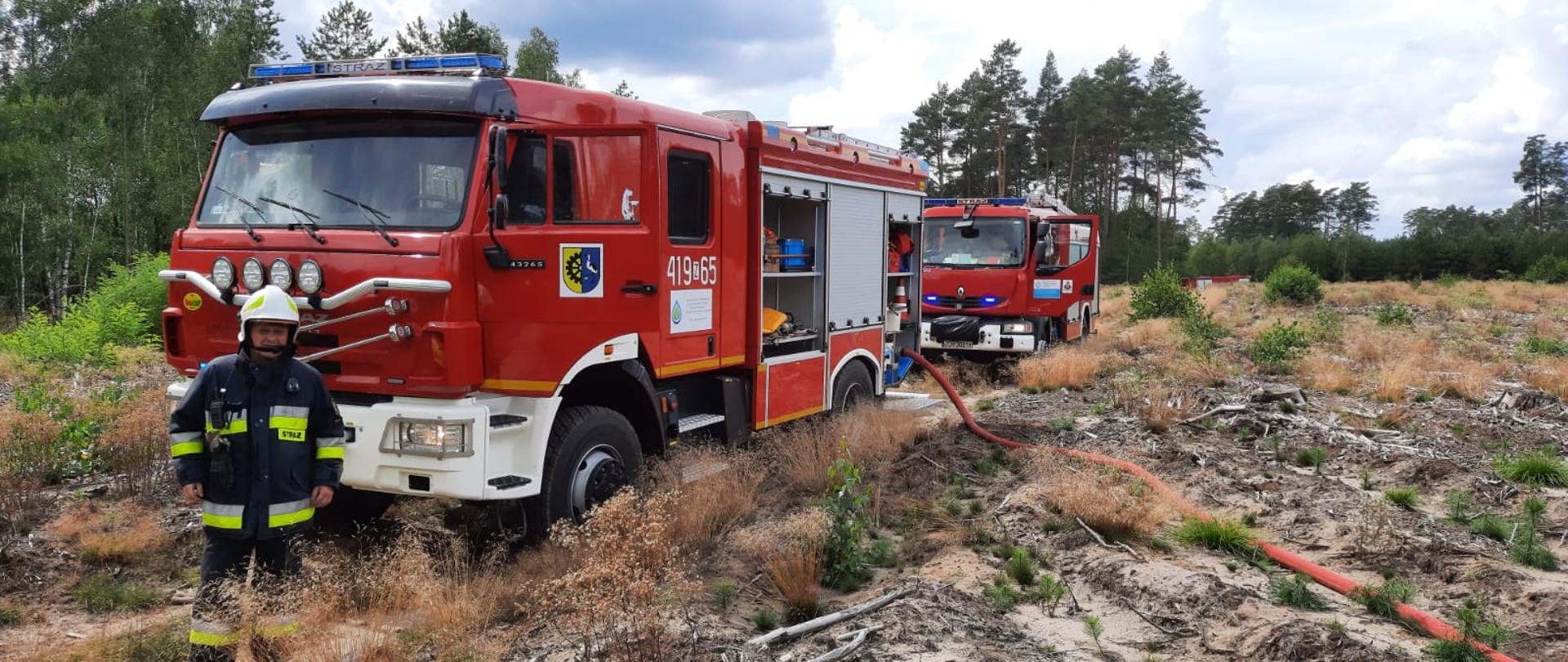 Na fotografii znajdują się dwa wozy strażackie, które mają otwarte drzwi od strony kierowcy. Samochody stoją na drodze leśnej. Dookoła są drzewa oraz krzaki. Przed pierwszym wozem stoi strażak ochotnik, który jest ubrany w ubranie specjalne. Z pierwszego wozu poprowadzony jest wąż gaśniczy, który znika w prawej części fotografii. W oddali widać trzeci wóz strażacki. Zdjęcie wykonano w porze dziennej. 