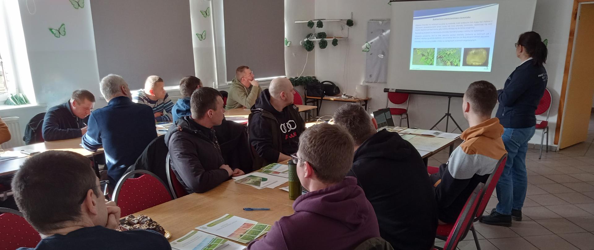 od lewej kobieta stoi zwrócona do wyświetlanego slajdu, przed nią w dwóch rzędach stołów siedzą mężczyźni 