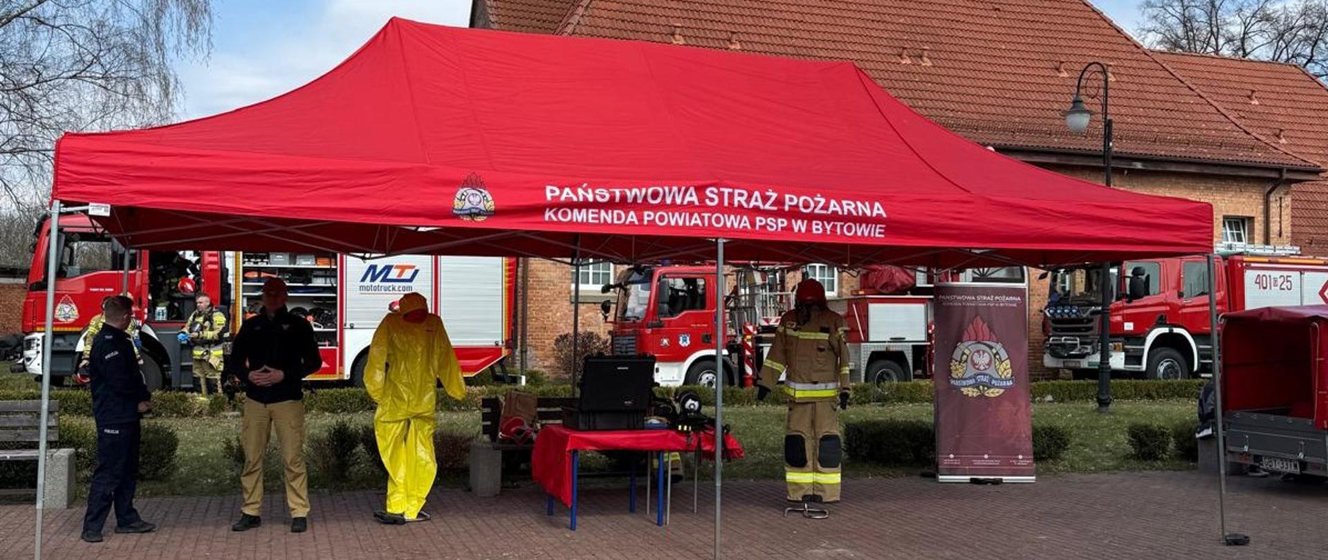 Namiot, bez ścian bocznych, z czerwonym dachem. Pod namiotem są funkcjonariusz PSP i Policji. Oprócz tego stoi mundur strażacki oraz na czerwonym stole jest sprzęt strażacki