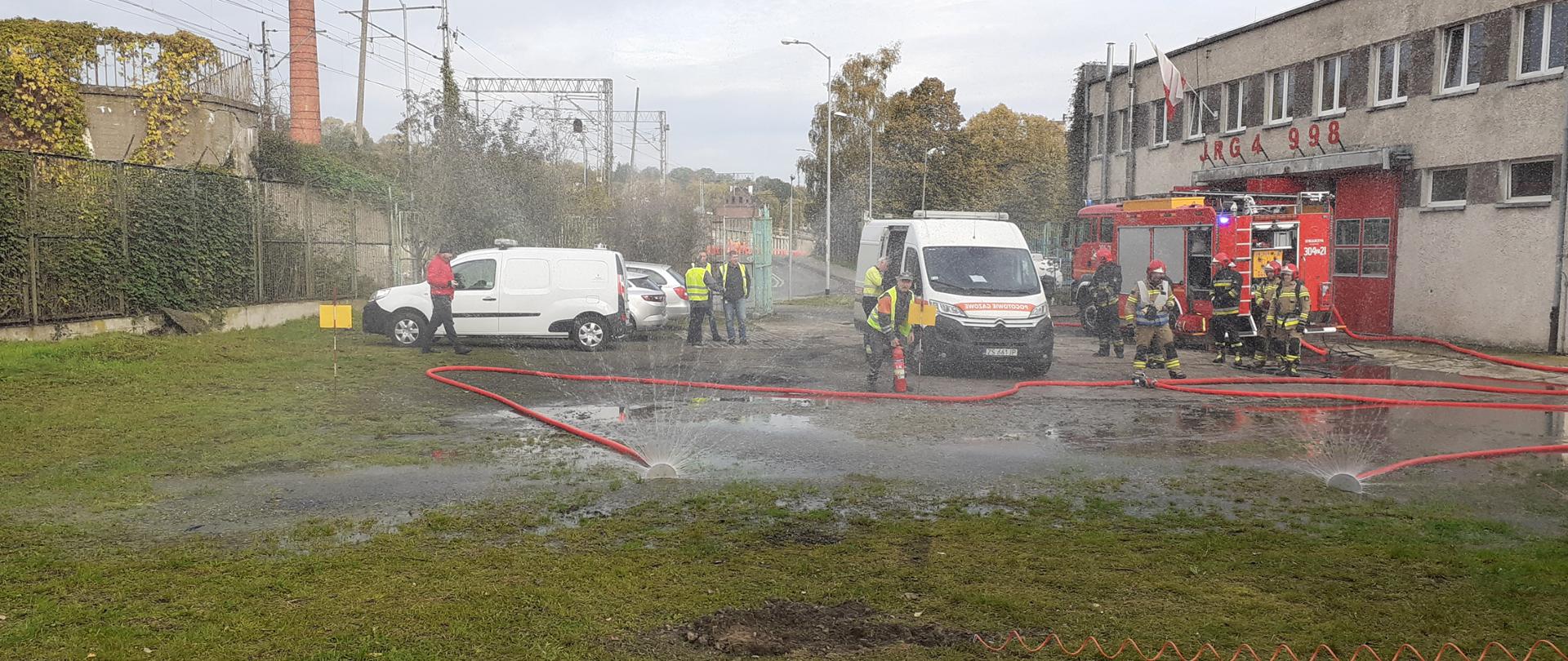 za dwoma rozstawionymi kurtynami wodnymi widać pracowników Pogotowia gazowego oraz ich samochód typu bus. obok widać samochód strażacki oraz strażaków czekających na rozkazy.