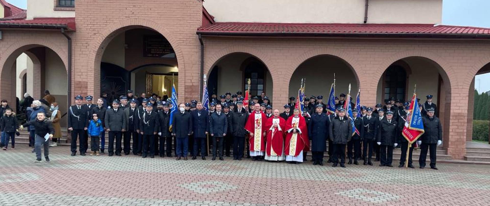 Zdjęcie przedstawia pozujących do zdjęcia grupowego strażaków z powiatu zambrowskiego ubranych w mundury wyjściowe ze sznurem galowym, część strażaków dodatkowo w strażackich kurtkach zimowych. Na zdjęciu poza strażakami pozują również przedstawicie władz oraz księża ustawieni w centralnej części zdjęcia. Zdjęcie wykonano w pochmurny, jesienny dzień na placu przed wejściem do Kościoła Rzymskokatolickiego pod wezwaniem świętego Józefa Rzemieślnika w Zambrowie. W tle widać część budynku kościoła.