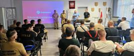 Na zdjęciu widoczna jest sala szkoleniowa, w której odbywa się spotkanie dla strażaków. Uczestnicy siedzą na krzesłach ustawionych w rzędach, przodem do prowadzących. Większość z nich ubrana jest w koszulki lub mundury z napisem „STRAŻ” lub „Państwowa Straż Pożarna”.
Z przodu sali stoją cztery osoby – trzech mężczyzn i jedna osoba prowadząca prezentację. Jeden z nich przemawia do uczestników, pozostali stoją obok. Za nimi znajduje się ekran projekcyjny, na którym wyświetlona jest prezentacja z napisem „Szkolenie z systemów fotowoltaicznych” oraz logo firmy Solplanet.
Sala jest jasna, z oświetleniem sufitowym i oknami po prawej stronie. Na ścianie za prowadzącymi widoczny jest godło Polski oraz flagi. Uczestnicy skupieni są na prezentacji, co wskazuje na formalny charakter szkolenia – prawdopodobnie dotyczącego bezpieczeństwa i działań ratowniczych związanych z instalacjami fotowoltaicznymi.