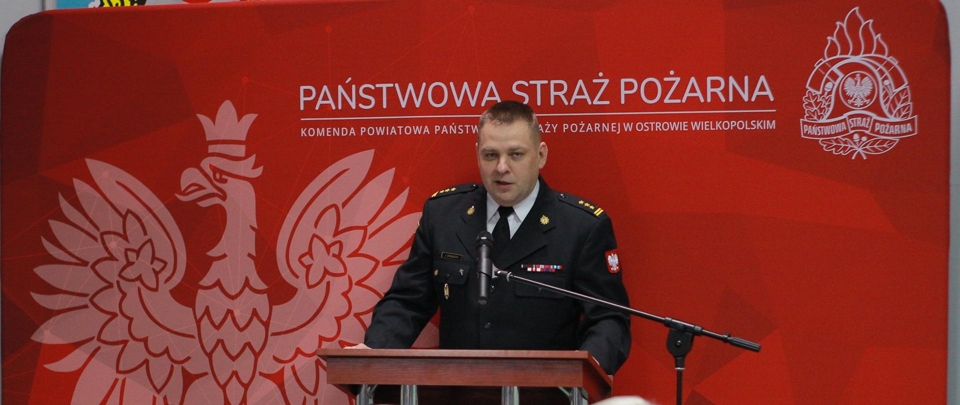 strażak stoi przed mównicą, przemawia. przed nim siedzą mężczyźni przy stole, za nim czerwona ścianka z orlem i napisem państwowa straż pożarna