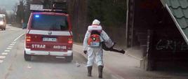 Na zdjęciu widoczny strażak w ubraniu ochronnym koloru białego w maseczce na plecach opryskiwacz spalinowy dezynfekuje wiatę przystankową środkiem odkażającym. Wiata drewniana pokryta blachą koloru zielonego. Przed strażakiem na drodze znajduje się samochód ratowniczo-gaśniczy koloru czerwonego.