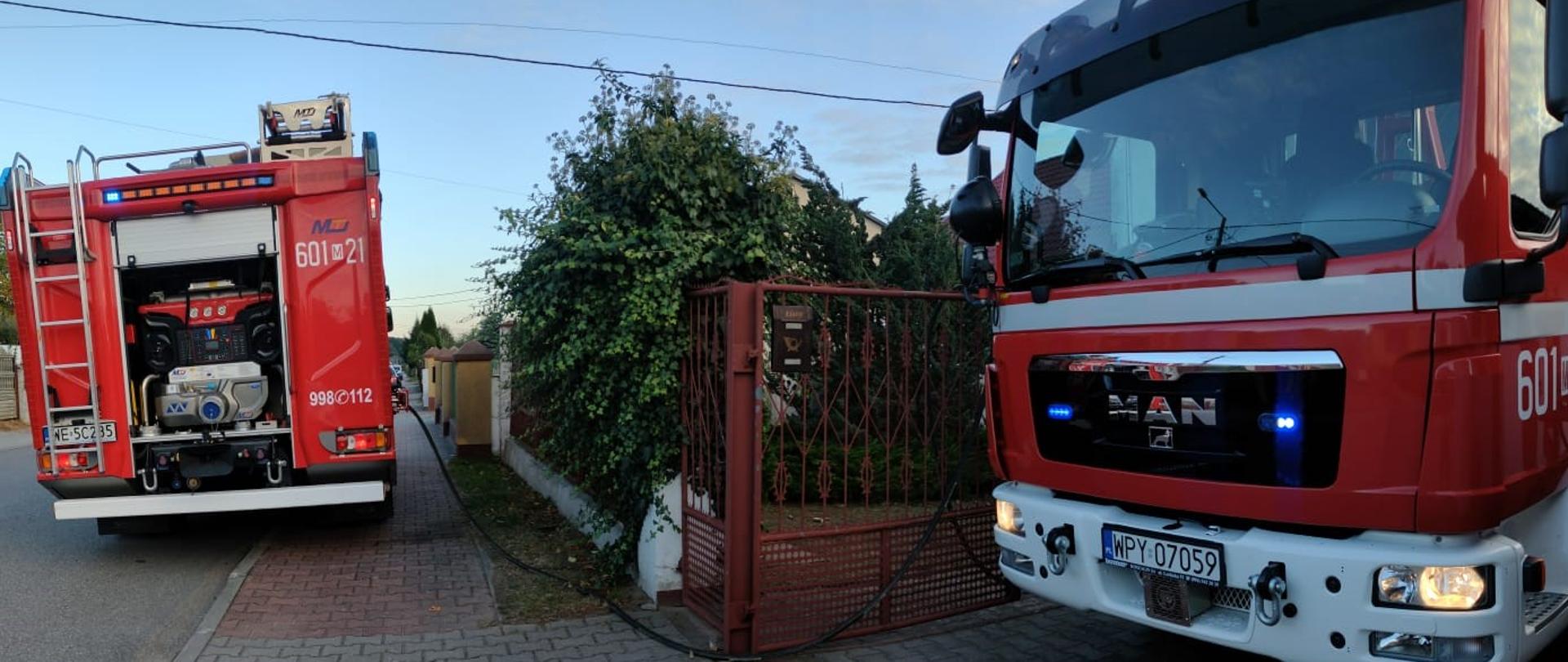 Zdjęcie przedstawia dwa czerwone wozy strażackie zaparkowane na wybrukowanej drodze przed posesją mieszkalną. Scena rozgrywa się o zmierzchu lub wczesnym rankiem, co widać po kolorze nieba. Na pierwszym planie, po prawej stronie, widać przód dużego wozu strażackiego z napisem "MAN" na masce i numerem "601 21" po prawej stronie kabiny. Włączone są niebieskie światła awaryjne pod przednią szybą. Samochód jest skierowany przodem w stronę widza. Z przodu pojazdu widać biały zderzak i włączone światła. Na dalszym planie, po lewej stronie, stoi tył drugiego wozu strażackiego z numerem "601 21" oraz numerami alarmowymi "998/112". Z tyłu wozu widać odsłonięty sprzęt gaśniczy i agregat prądotwórczy. Między wozami, na ziemi, leżą węże pożarnicze, co wskazuje na trwającą akcję lub jej zakończenie. Pomiędzy pojazdami widoczna jest brązowa brama wjazdowa do posesji, otoczona gęstymi krzewami. Po prawej stronie drogi, za bramą, widać fragment domu i drzewa. Cała scena sugeruje interwencję straży pożarnej na terenie prywatnej posesji.