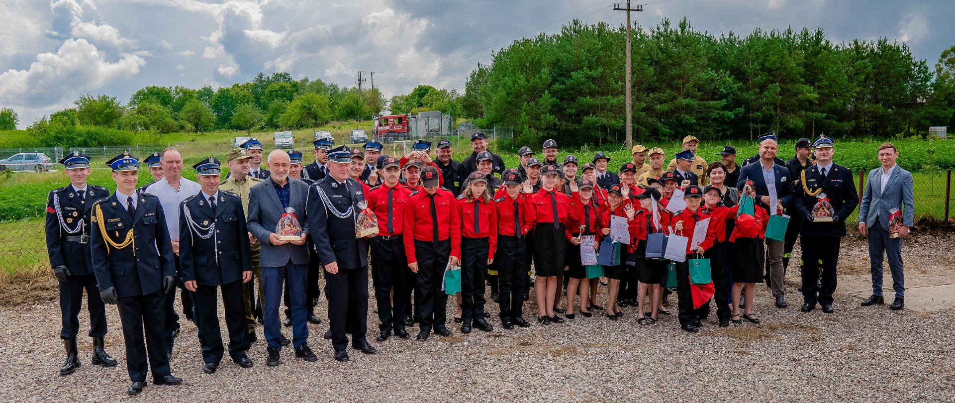 Zdjęcie przedstawia uczestników uroczystego apelu z okazji ślubowania nowych członków Młodzieżowej Drużyny Pożarniczej oraz druhów Ochotniczych Straży Pożarnych w Surminach, zlokalizowanych na placu przy strażnicy