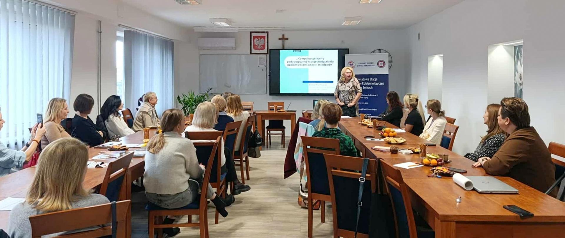 Kobieta prowadzi prelekcję na temat kompetencji kadry pedagogicznej dla grupy kilkunastu uczestników siedzących przy długich stołach w sali konferencyjnej. Na ekranie za prelegentką wyświetlana jest prezentacja.