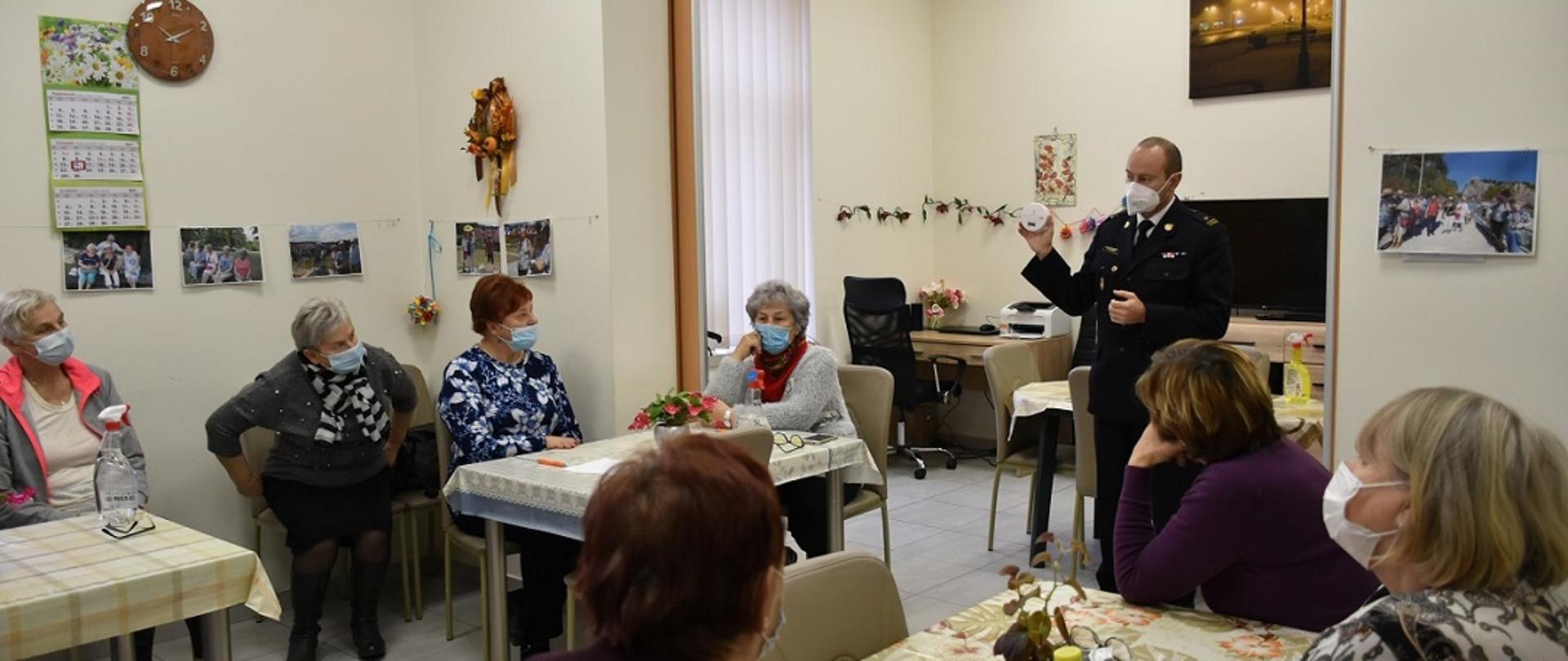 W tle zdjęcia znajduje się funkcjonariusz Komendy Powiatowej PSP w Skarżysku-Kamiennej, który w ręce trzyma czujnik czadu. Jest on w trakcie prowadzenia pogadanki z zakresu prawidłowego przygotowania budynków mieszkalnych do trwającego sezonu grzewczego. Pogadanka jest przeprowadzana dla seniorów z klubu seniora znajdującego się na terenie powiatu skarżyskiego. Ze względu na trwającą pandemię wszyscy uczestnicy spotkania mają założone maseczki na usta i nos. 
