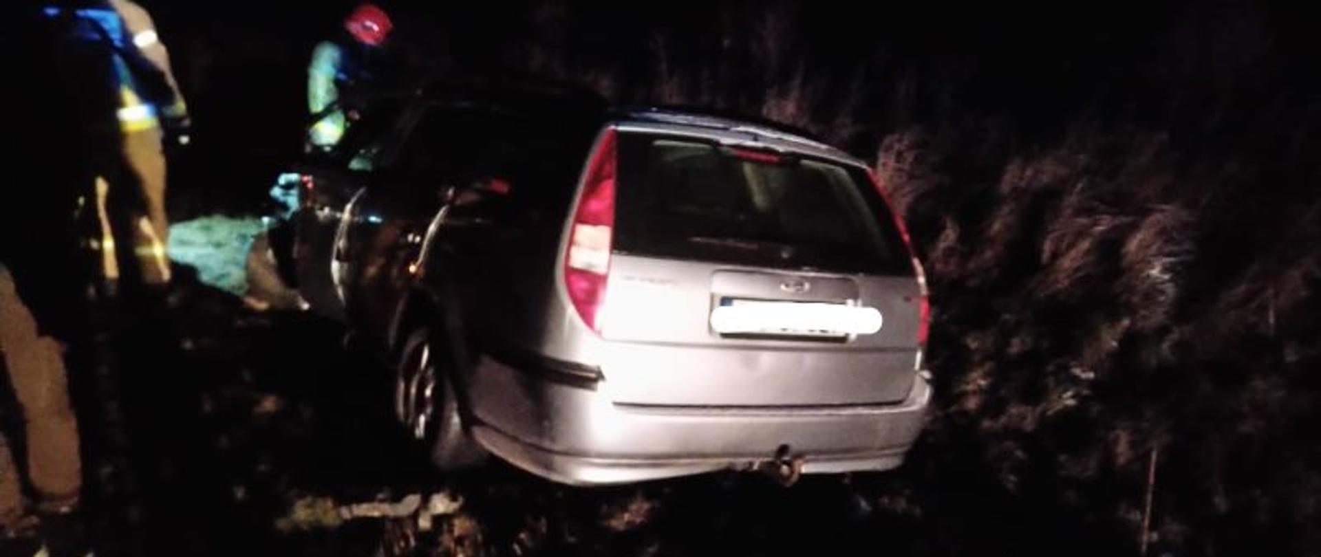 Samochód osobowy w wersji kombi po uderzeniu w drzewo. Pojazd oświetlony lampami najaśnic strażackich. W oddali widać strażaka w piaskowym mundurze, który prowadzi czynności