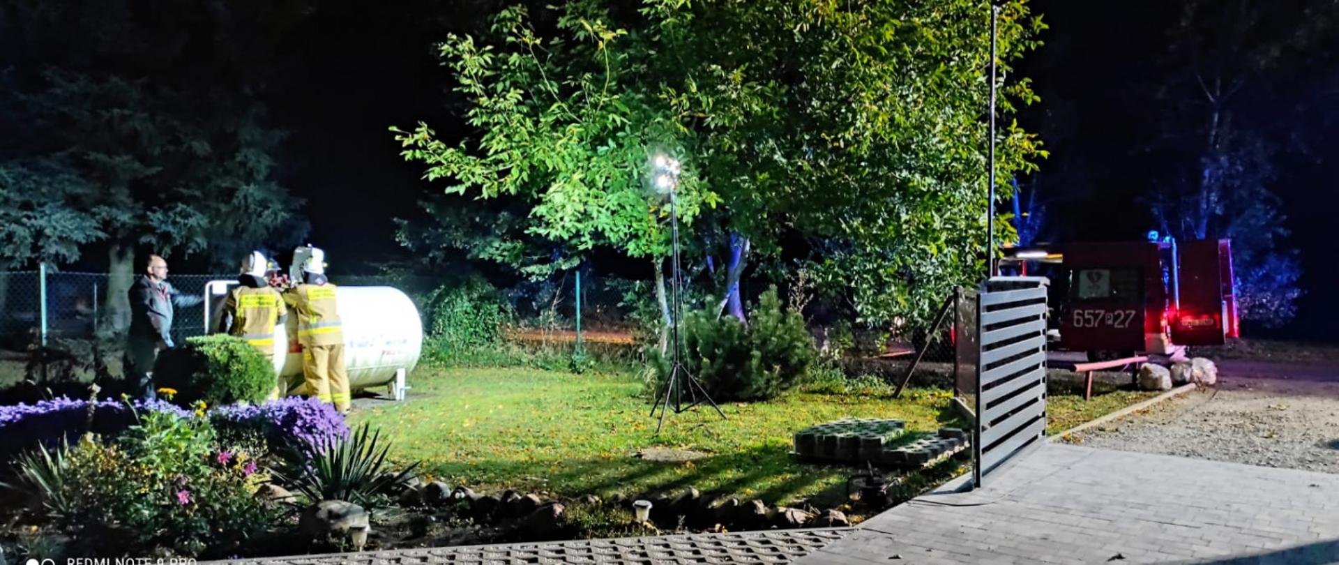 Zdjęcie przedstawia strażaków stojących przy zbiorniku z gazem. Obok wóz strażacki i oświetlenie terenu akcji.