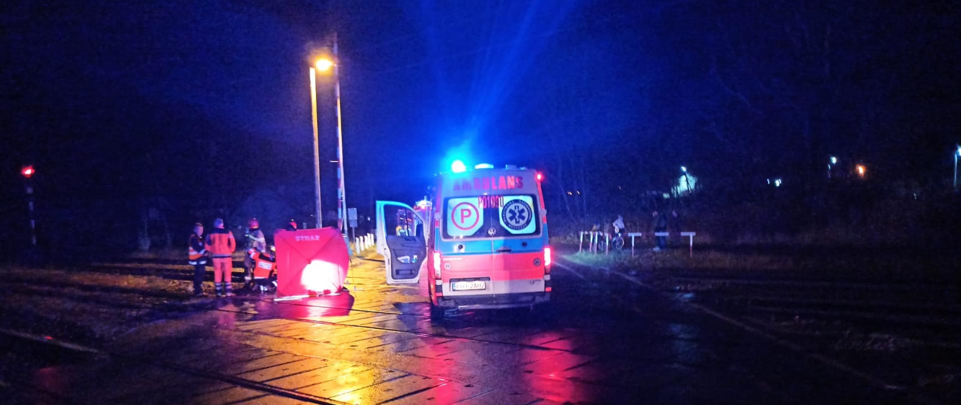 Zdjęcie wykonane na przejeździe kolejowym. Na środku stoi ambulans z włączonymi światłami alarmowymi. Po jego lewej stronie rozłożony jest parawan ratowniczy, a obok niego stoją strażacy i ratownicy.