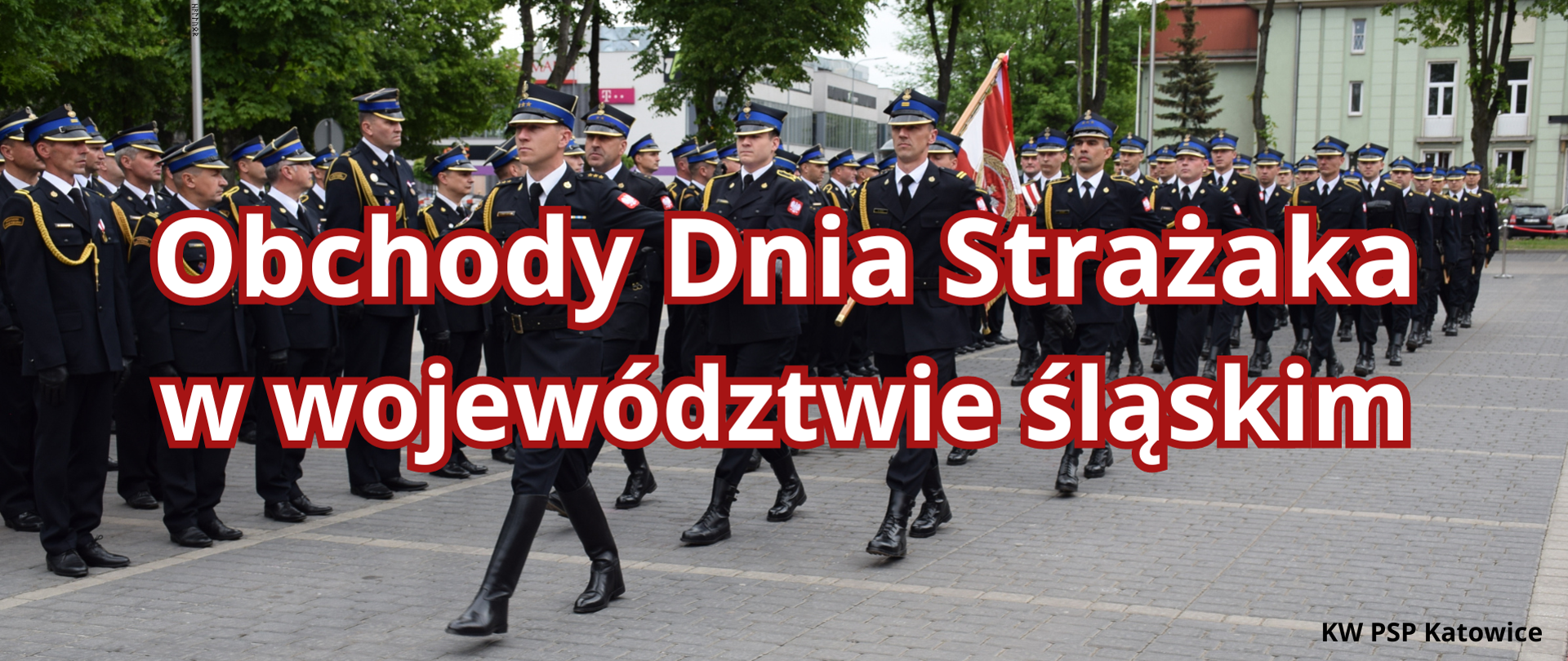 na zdjęciu widoczni strażacy w mundurach wyjściowych z komapanii reprezentacyjnej Komendy Wojewódzkiej Państwowej Straży Pożarnej w Katowicach. Strażacy maszeruja w szyku. Jako pierwszy dowódca kompanii za nim strażacy z pocztu sztandarowego. Na zdjęciu widnieje napis w kolorze białym z czerwonym obramowaniem Obchody Dnia Strażaka w województwie śląskim