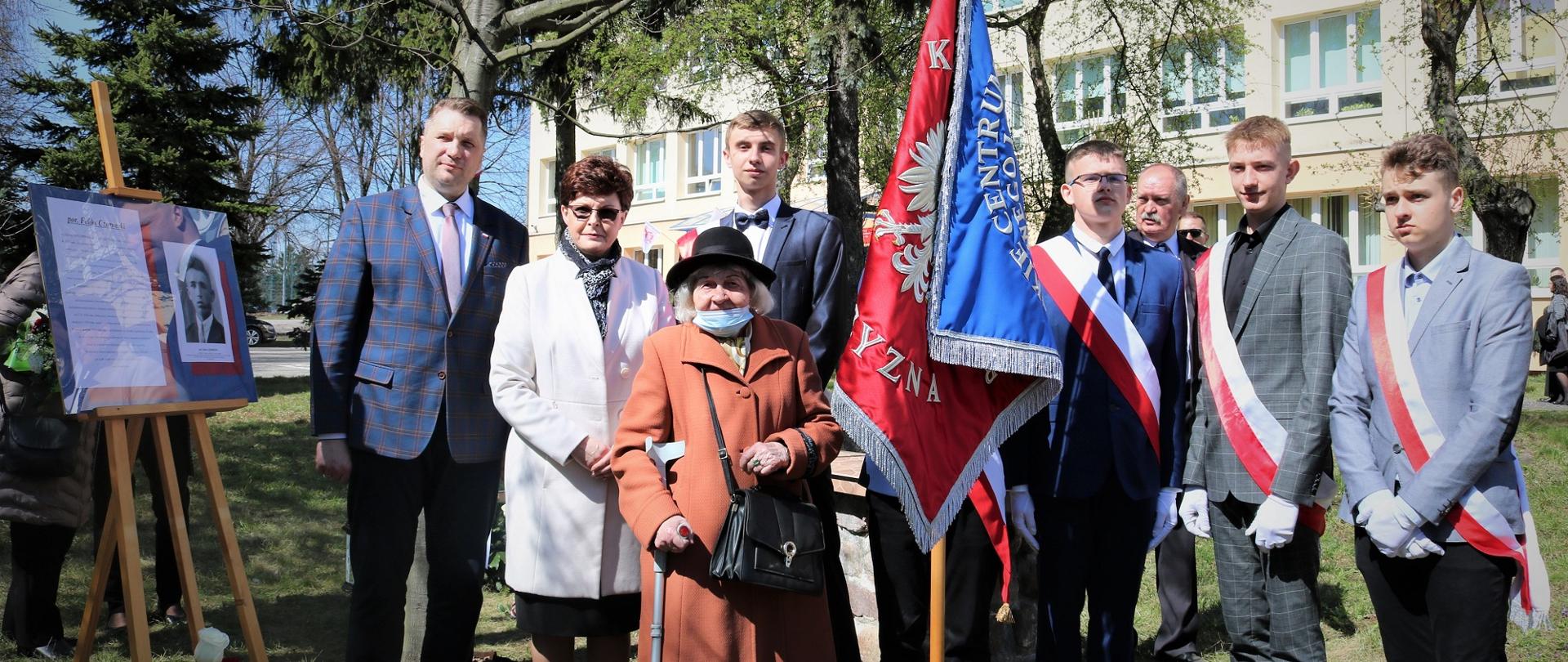 Minister stoi przed budynkiem szkoły w towarzystwie grupy elegancko ubranych osób, przed nimi sztandar.
