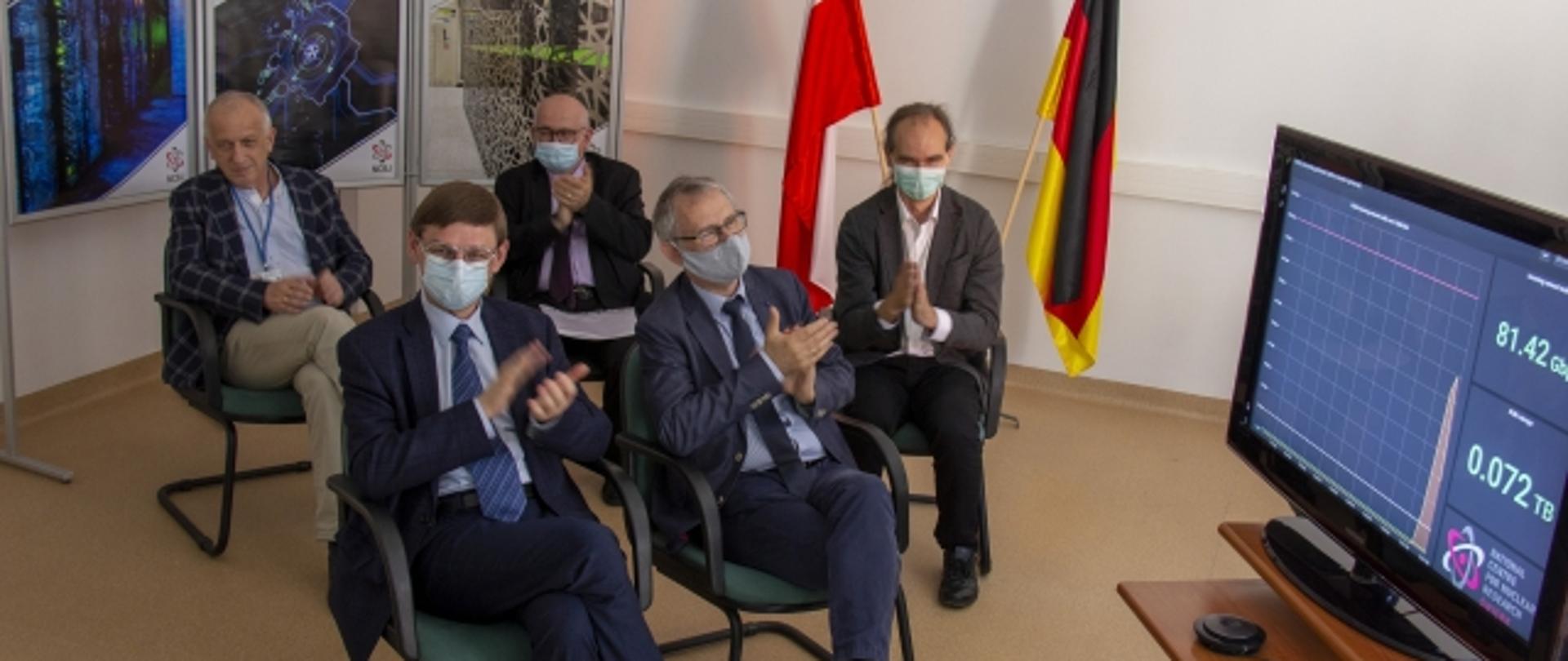 Zdjęcie przedstawiające grupę 5 mężczyzn siedzących przed dużym monitorem, w tle widoczne są dwie flagi - polska i niemiecka 