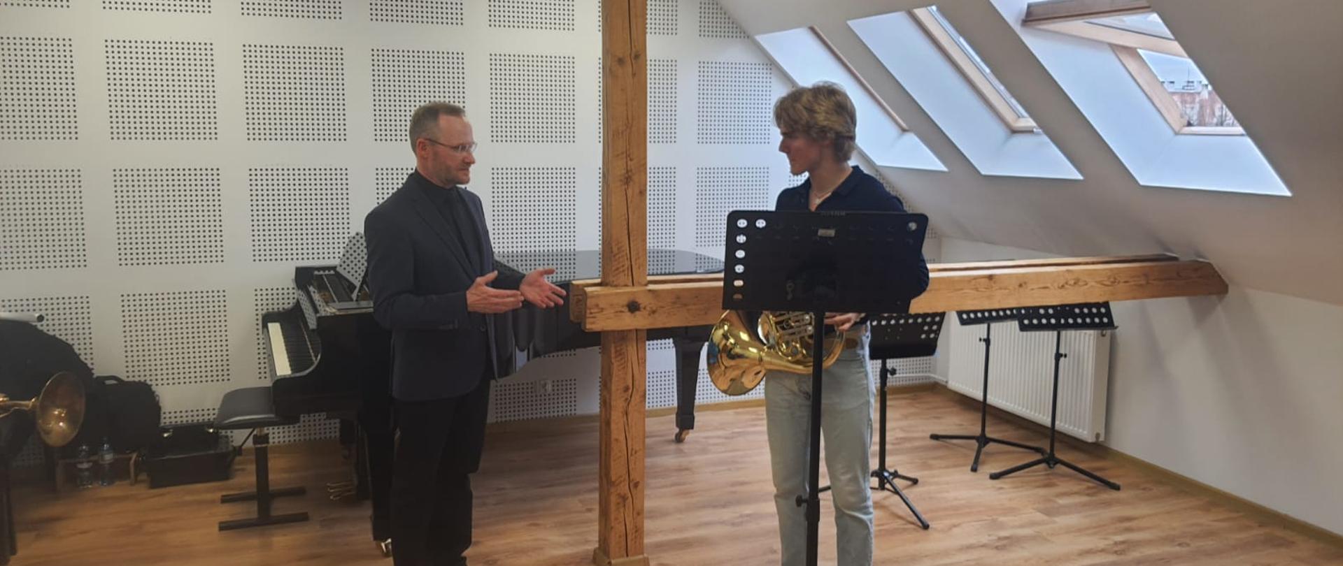 na zdjęciu widać ucznia który gran na puzonie obok niego stoi nauczyciel przy fortepianie przed uczniem stoi pulpit z nutami w czarnym kolorze