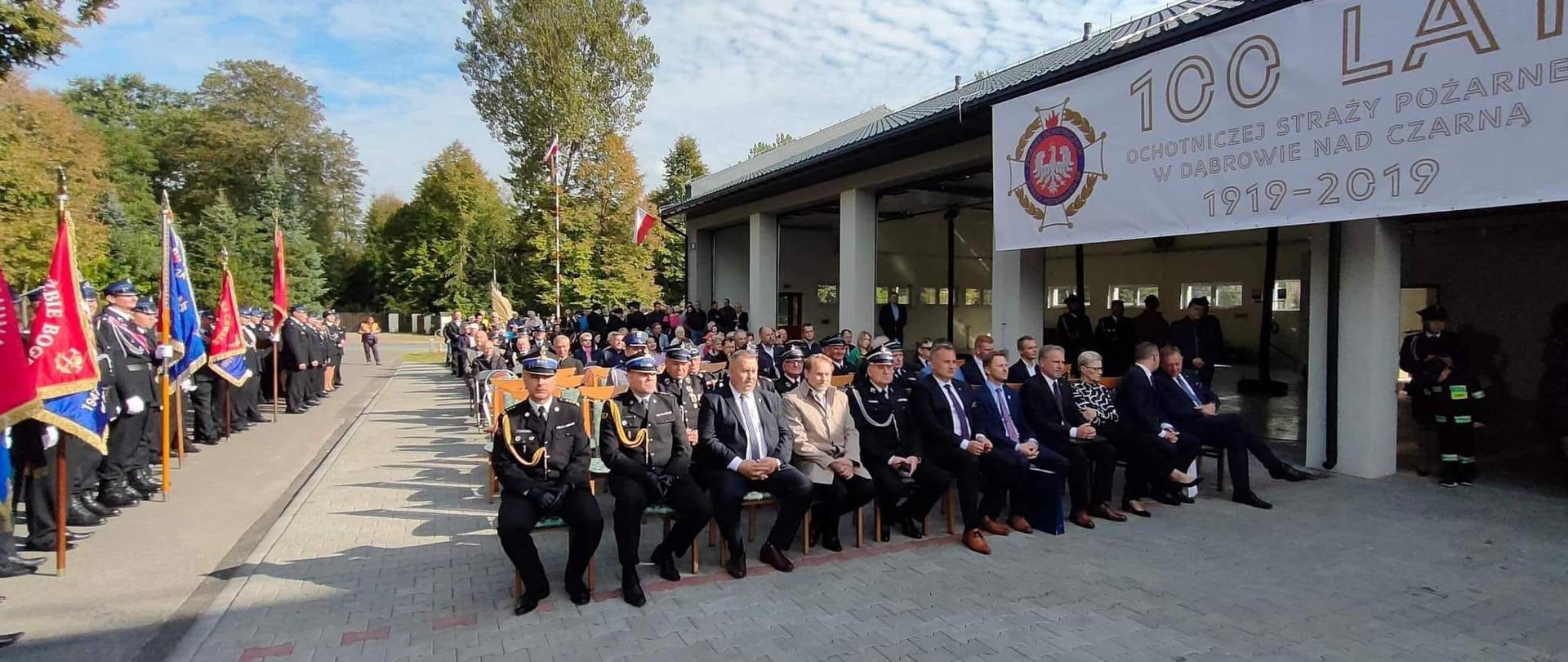 strażacy i inni ludzie siedzący na krzesłach przed garażami jednostki osp po lewej stronie sztandary osp
