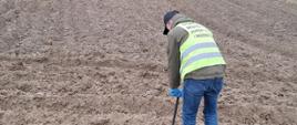 Na polu inspektor WIORiN w Kielcach z Oddziału w Opatowie pobiera próby gleby na obecność mątwików (Globodera pallida i Globodera rostochiensis) metalową laską glebową.
Inspektor ubrany jest w niebieskie spodnie i zieloną kurtkę na którą nałożoną ma odblaskową kamizelkę z napisem Państwowa Inspekcja Ochrony Roślin I Nasiennictwa, na jego butach i rękach widoczna jest odzież ochronna (ochraniacze foliowe na buty i rękawiczki jednorazowe koloru niebieskiego).
