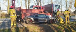 Kolizja drogowa-dachowanie audi. Droga gminna Rawicz-Przywsie. Na tle samochodu ciężarowego znajduje się rozbite audi. Trwają przygotowania do załadunku. Wokół audi znajduje się 4 strażaków. W tle drzewa.